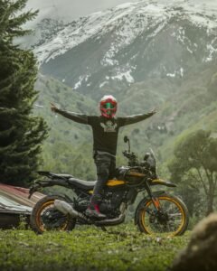 Motorcyclist with arms spread celebrates journey against snowcapped mountains.